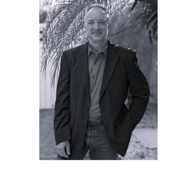 Man in a blazer standing outdoors with a relaxed smile.