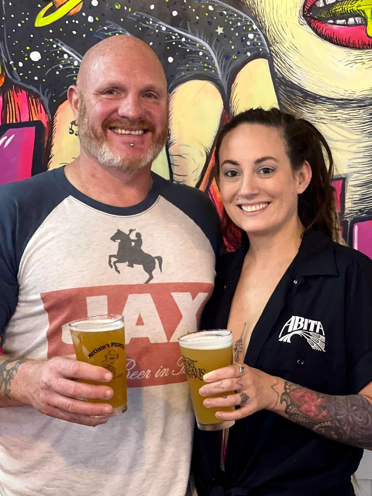 Two people smiling with beers in hand, standing in front of a colorful mural.