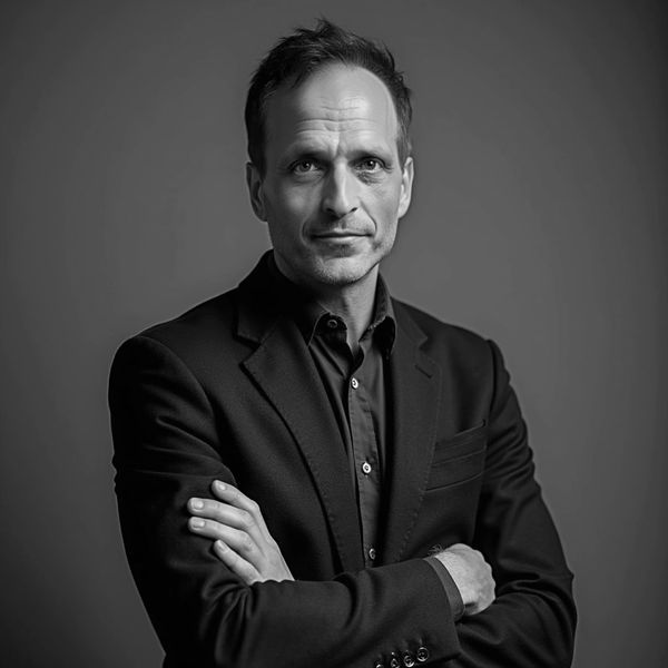 Confident man in black suit with arms crossed against gray backdrop.