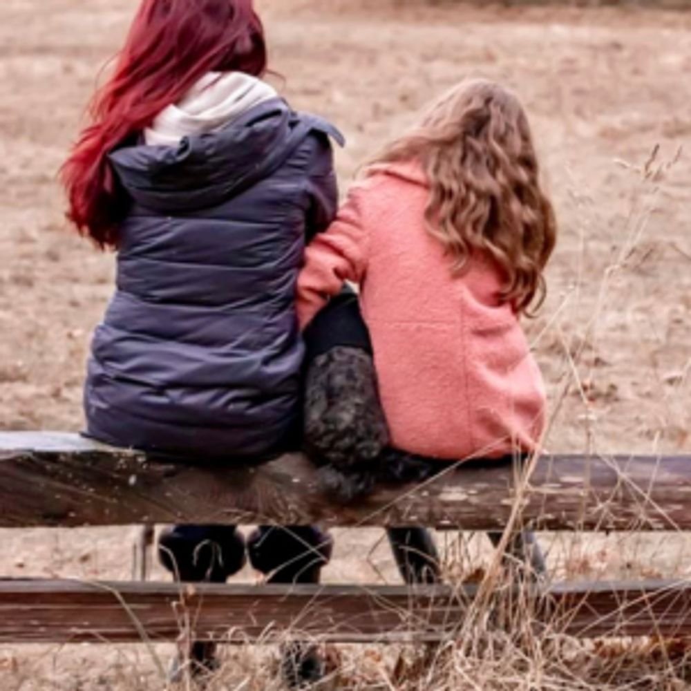 Two girls sitting on a bench with a dog between them, outdoors.