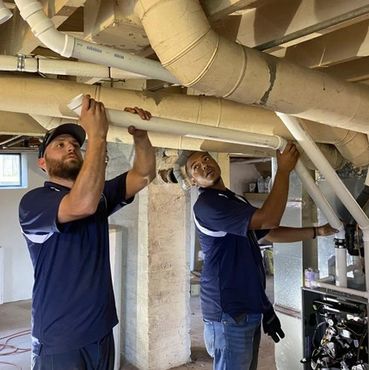Two workers installing pipes in a basement ceiling.