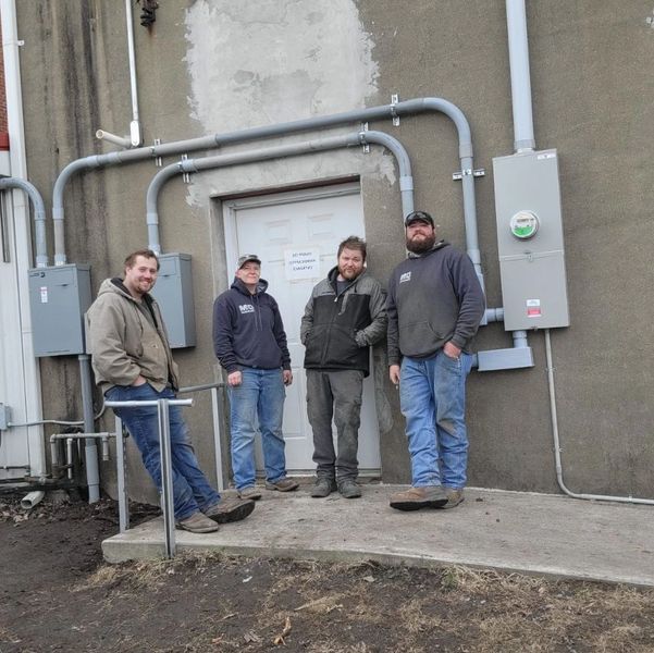 Four men standing casually outside a building with utility pipes and meters.