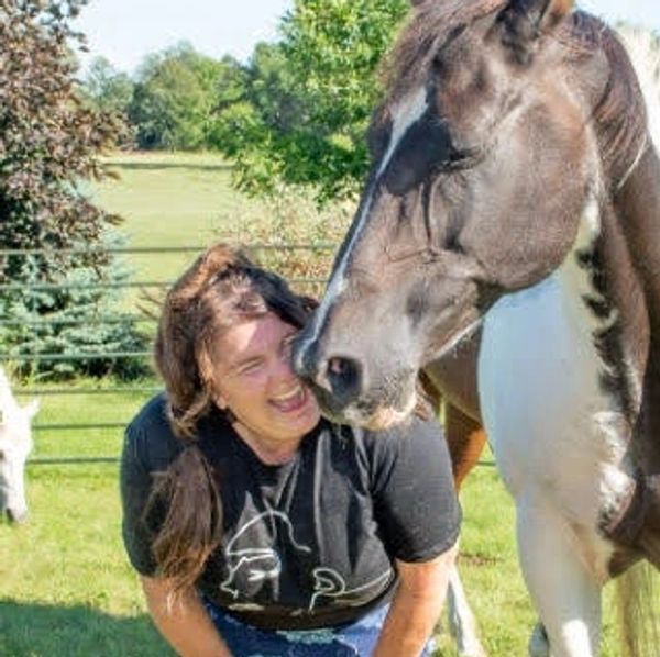A woman laughing as a horse nuzzles her face in a sunny field.