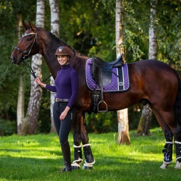 A woman in equestrian gear standing beside a saddled horse in a forest clearing.