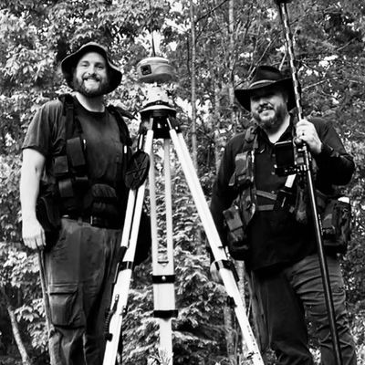 Two men with hats and gear, standing outdoors with surveying equipment.