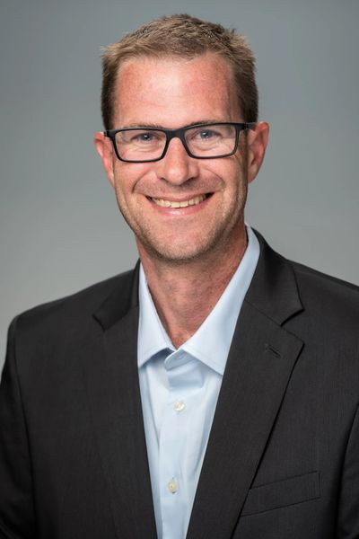 Smiling man in glasses wearing a black suit and light blue shirt.
