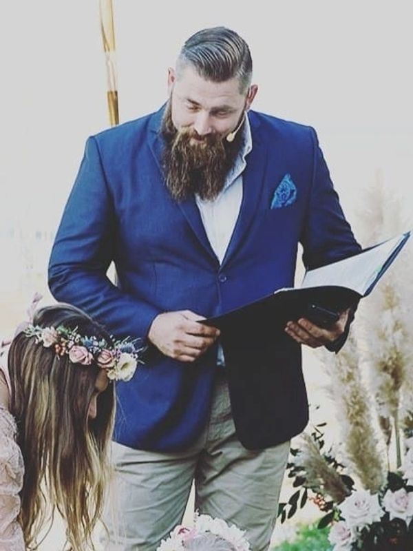 Man in blue blazer officiating a wedding with a bride wearing a floral crown.