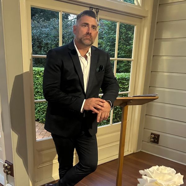 Man in a black suit leaning against a wooden podium indoors.