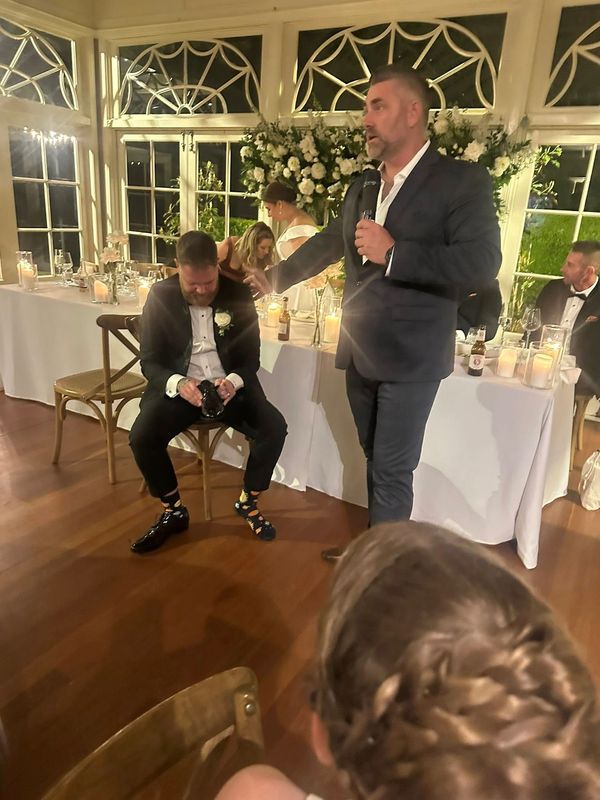 Man giving a speech while another man sits laughing at a wedding reception.