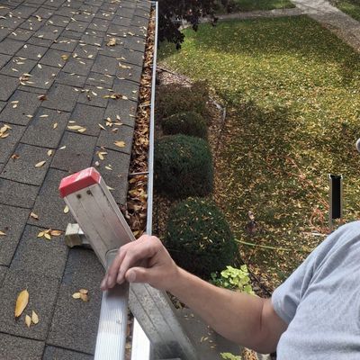 Person on ladder cleaning a leaf-filled gutter.