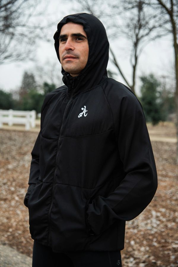 Man in a black hooded jacket stands outdoors on a cloudy day.