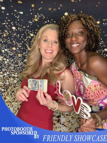 Two women smiling in a photobooth with props including money and a LOVE sign.