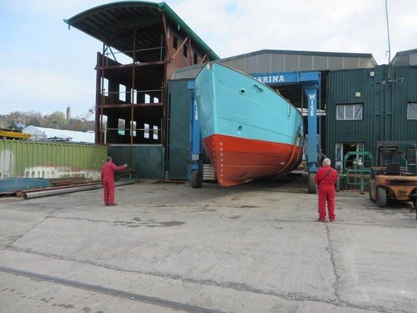 Workers inspecting a large boat hull outside a marina building.