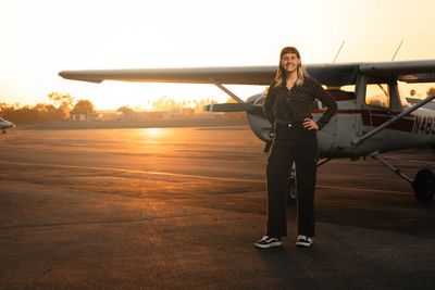 A woman standing confidently by a small airplane at sunset.