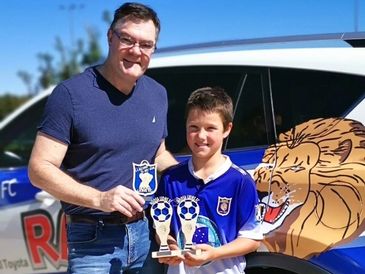 Man and boy holding soccer trophies in front of a lion-themed vehicle.