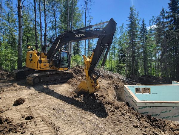 A yellow John Deere excavator digging near a construction site surrounded by trees.
