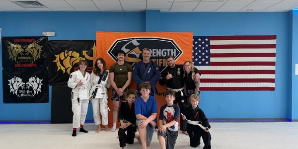 Group of martial arts students and instructors posing in front of flags inside a dojo.