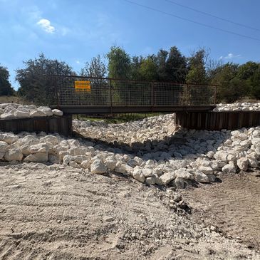 A small metal bridge over a rocky, dry drainage area in a rural setting.