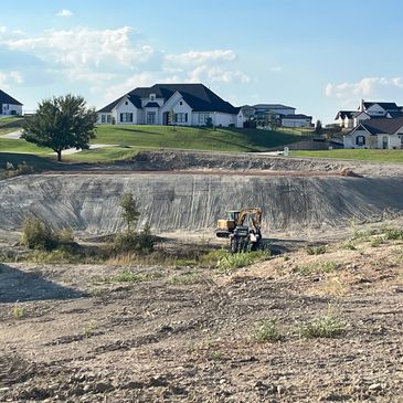 Construction equipment working in a large dug hole near suburban houses.
