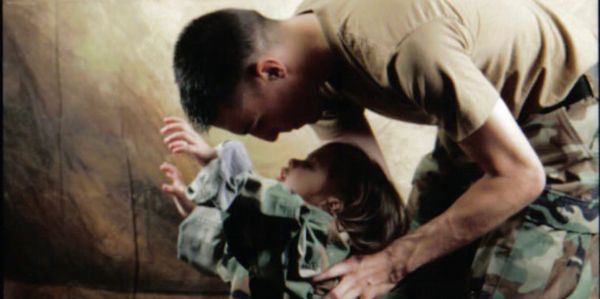 A soldier bending down to hug a child dressed in oversized camouflage. army brat