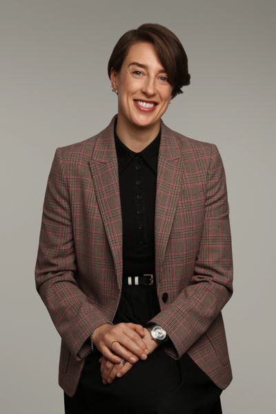 Smiling woman in a plaid blazer and black dress sits with hands clasped.