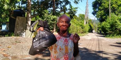 An elderly woman holding a black bag on a sunny rural road.