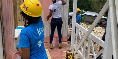 Two women in hard hats painting wooden walls outdoors.