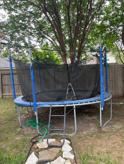 Backyard trampoline with safety net, ladder, and string lights under large trees.