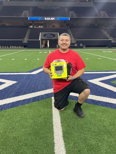 Man kneeling on a football field star, holding an AED device.