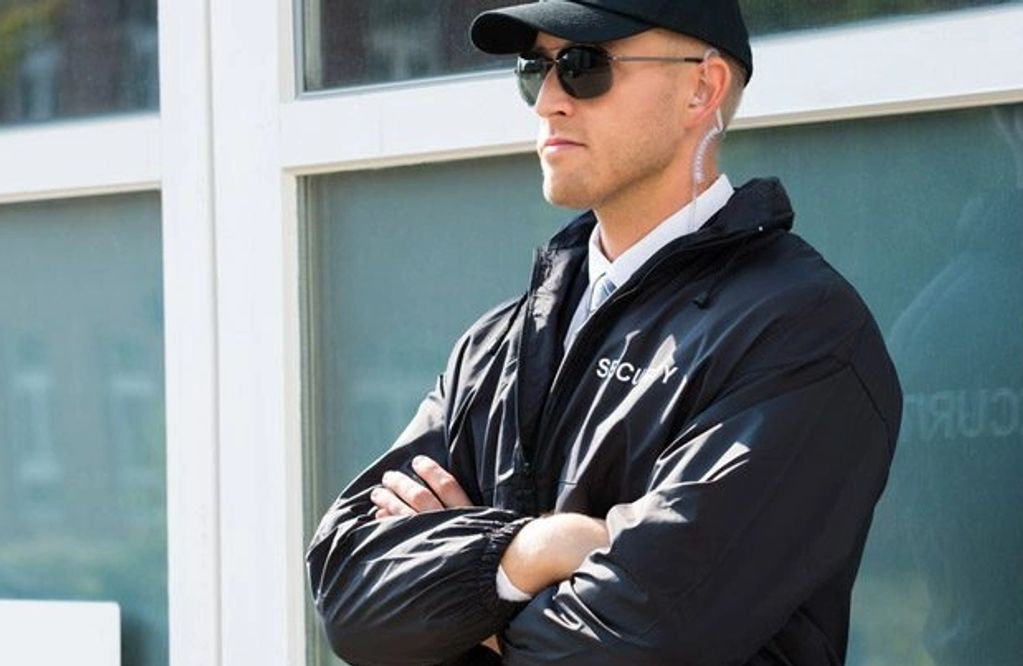 A security guard in sunglasses and uniform stands with arms crossed.