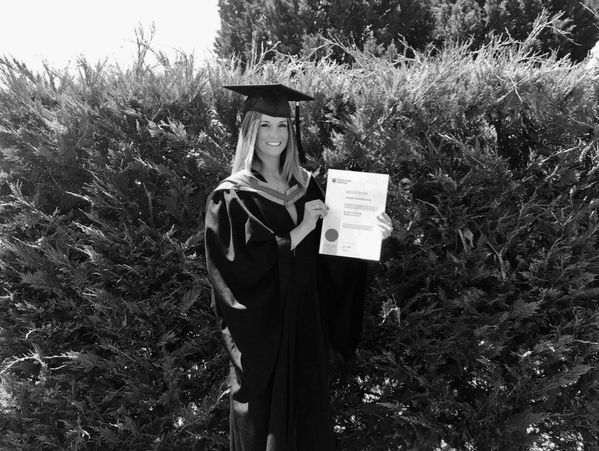 A smiling graduate in a cap and gown holding a diploma outdoors.