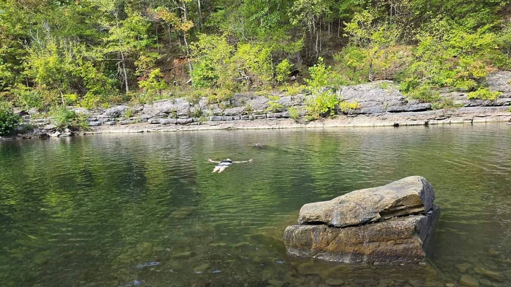 A person floating on their back in a calm river surrounded by green trees and rocks.