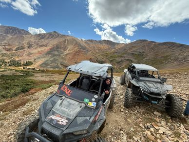 Two off-road vehicles on a rocky mountain trail under a partly cloudy sky.