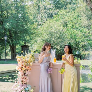 Two women in elegant dresses enjoying drinks by a flower-adorned bar outdoors.