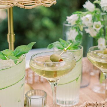 Elegant cocktails with olives and fresh herbs on a wicker tray outdoors.
