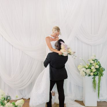 Groom lifts bride in a joyful wedding moment with elegant white drapery and floral arrangements.