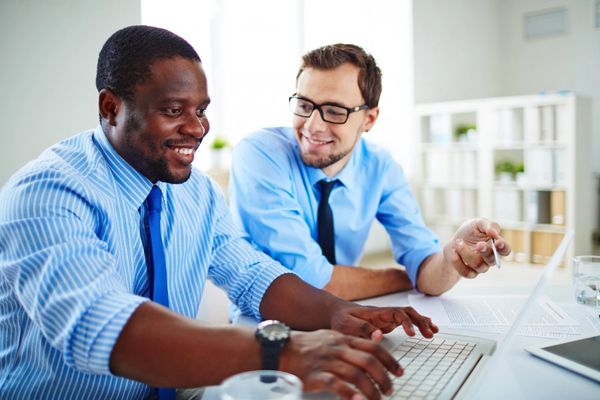 Two professionals in button-up shirts collaborate over a laptop on a desk filled