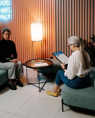 A woman with gray hair and glasses sits on a couch, reviewing documents while another person