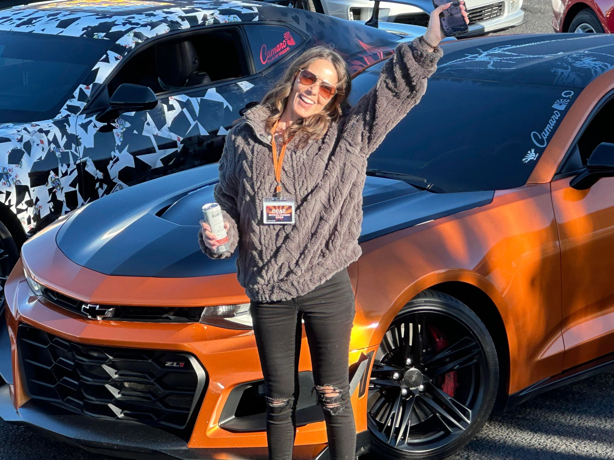 Woman smiling and posing in front of two Camaro ZL1 cars, holding a drink and phone.