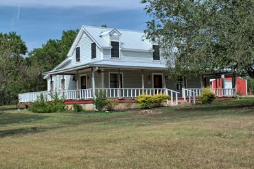 White country house with wraparound porch surrounded by greenery.