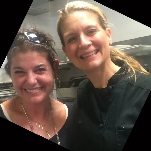 Two women smiling, one in a chef's coat, in a kitchen setting.