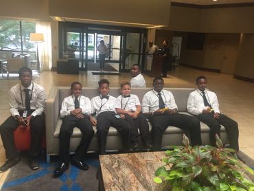 Seven young men in white shirts and ties sitting on a couch in a hotel lobby.