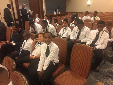 Young boys in white shirts and ties seated in an auditorium.