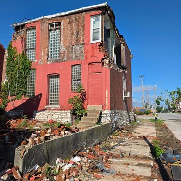 Partially collapsed brick building with debris on sidewalk under clear blue sky.