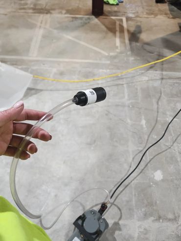 Hand holding a small black device attached to a transparent tube in an industrial setting.