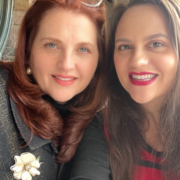 Two women smiling together indoors, one wearing glasses on her head.