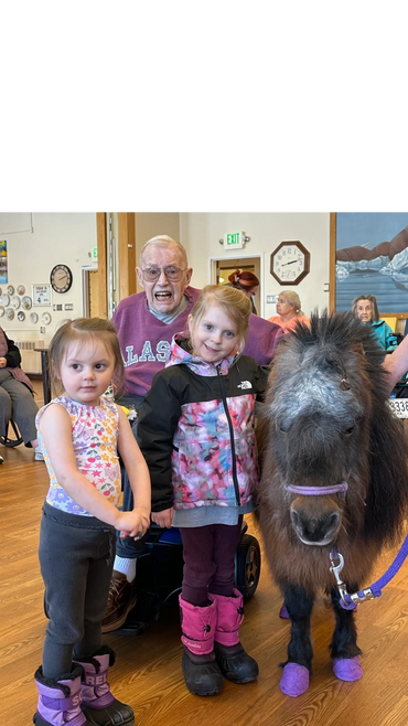 Two young girls with an elderly man and a miniature horse indoors.