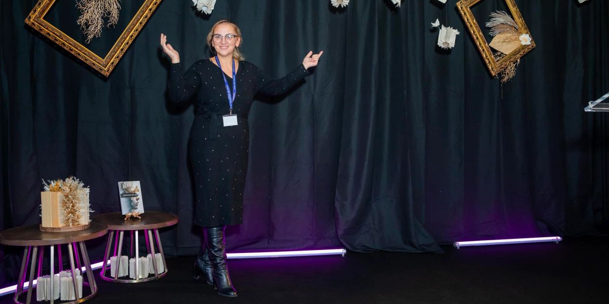 Woman in black dress posing with arms raised against a dark curtain backdrop.