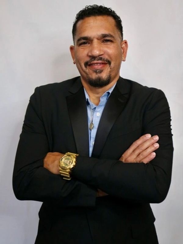 Man in black suit with gold watch smiling.