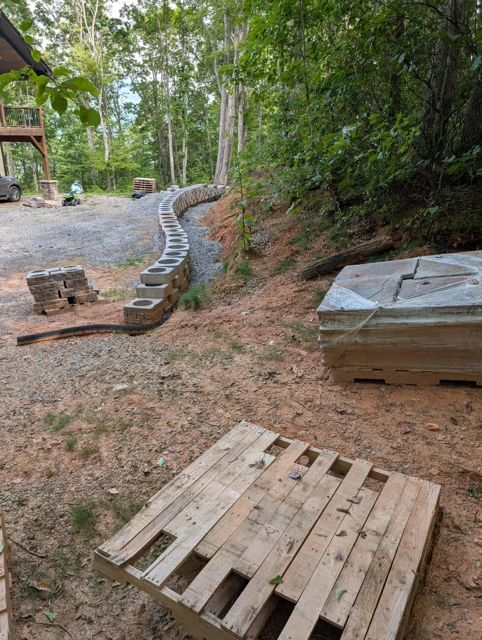 A backyard construction site with stacked bricks and wooden pallets near a wooded area.